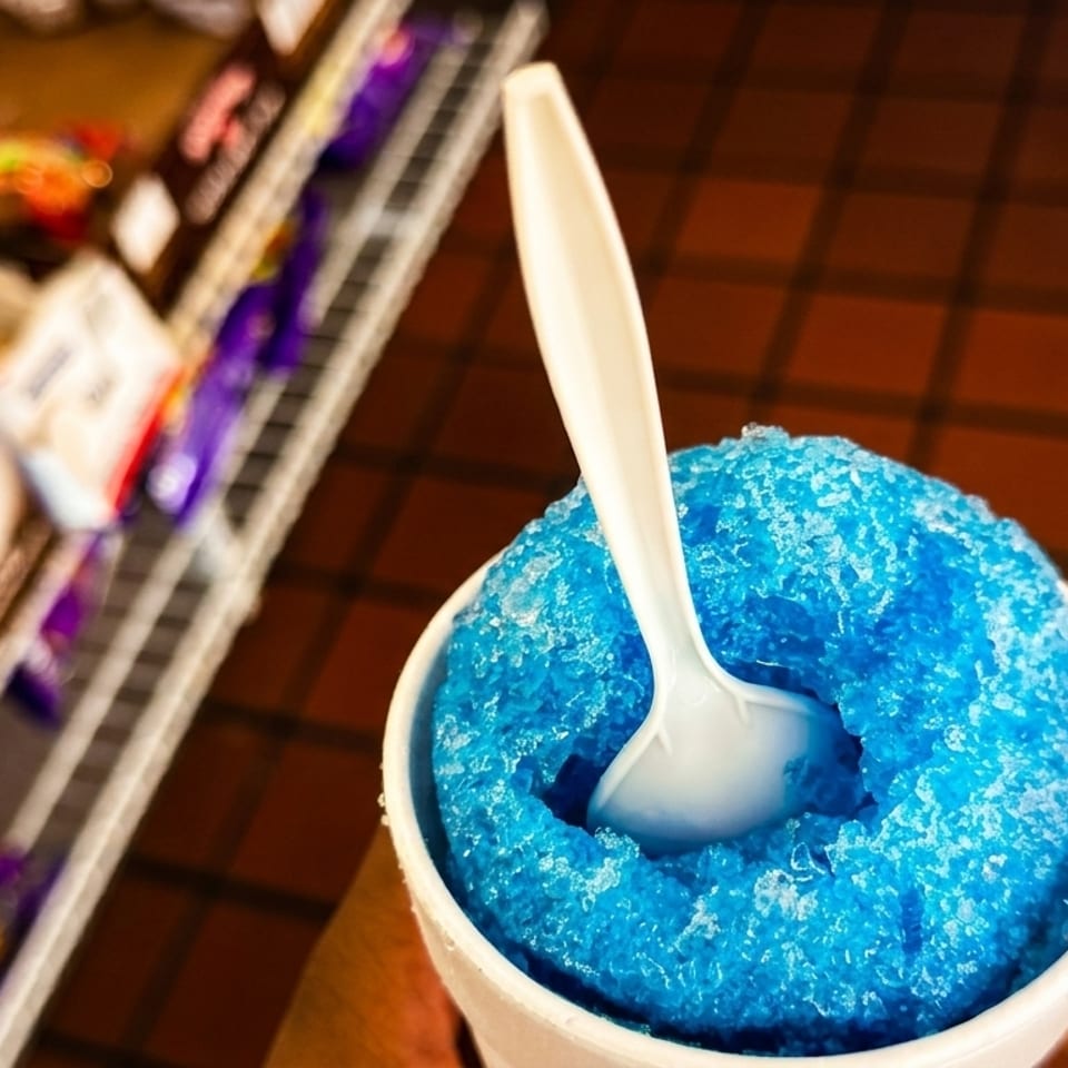 A bright blue shaved ice in a white cup with a spoon, against a blurred background of snacks.