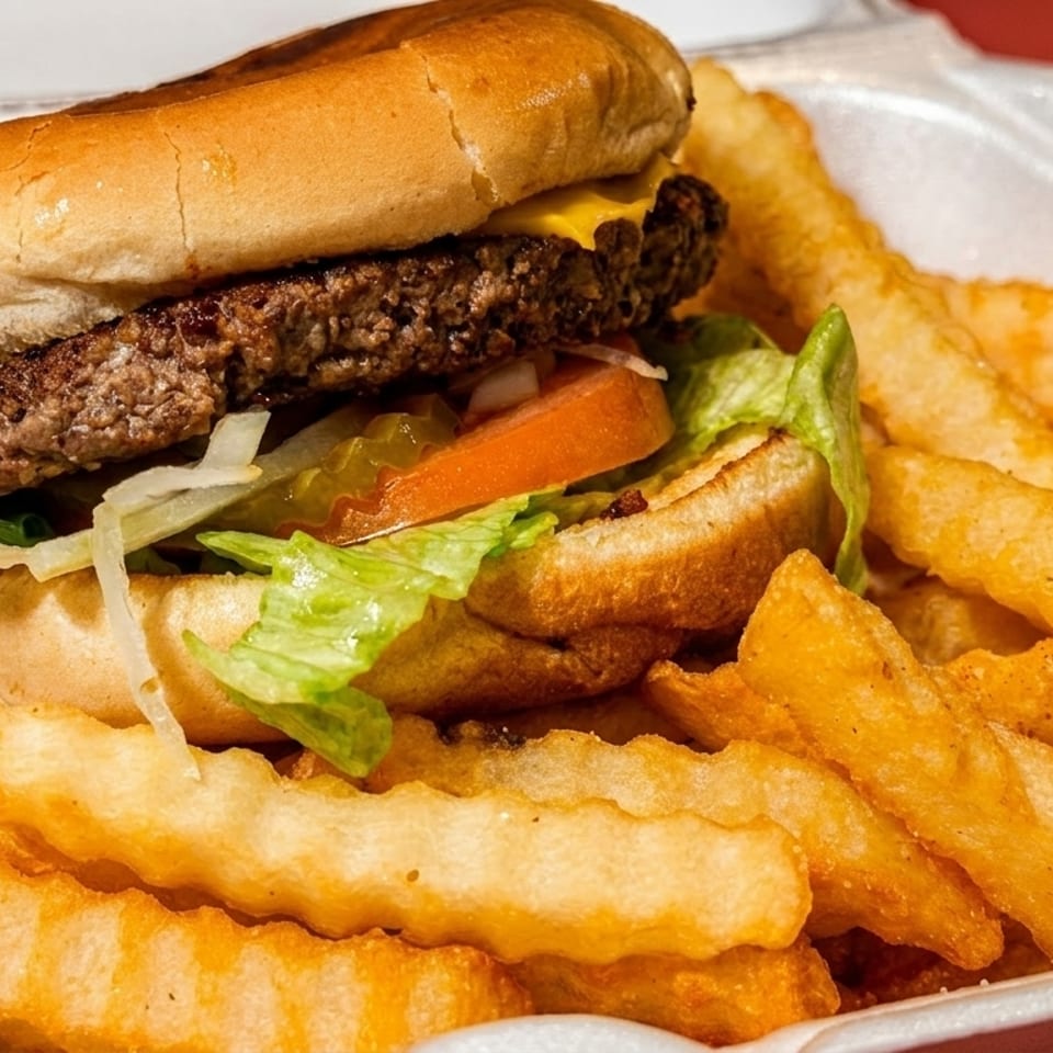 Cheeseburger with lettuce, tomato, and pickles, served with crinkle-cut fries in a white takeout container.